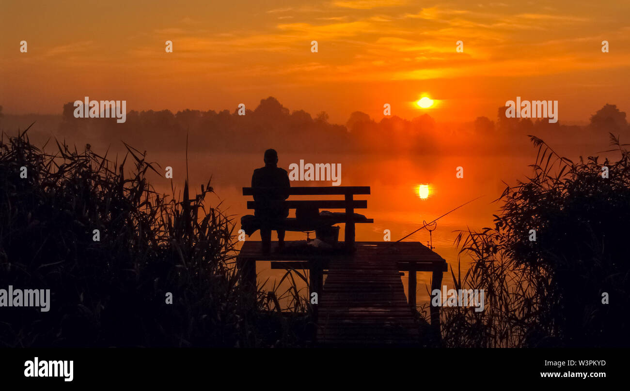 Angler seating on the wooden pier by the lake in the sunrise holding fishing rod. Silhouette landscape with beautiful summer sunlight. Stock Photo