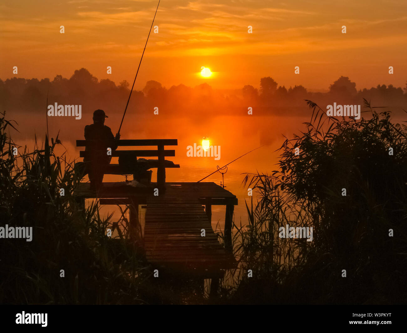 Angler seating on the wooden pier by the lake in the sunrise holding fishing rod. Silhouette landscape with beautiful summer sunlight. Stock Photo