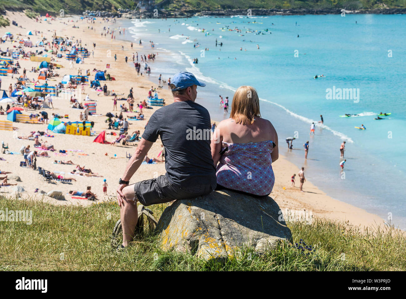 Holidaymakers sitting on a rock enjoying the sunny weather and the view over Fistral Beach and the turquoise coloured sea in Newquay in Cornwall. Stock Photo