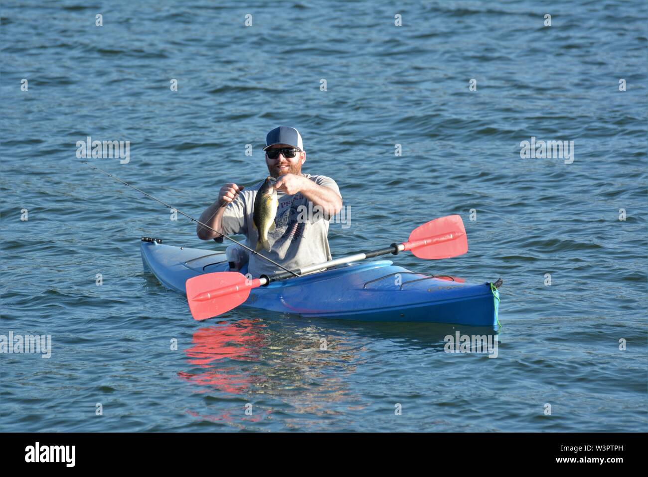 A real happy fisherman with caught small bass on his blue kayak on Clear Lake Clearlake California USA America on calm sunny day in summer by himself Stock Photo