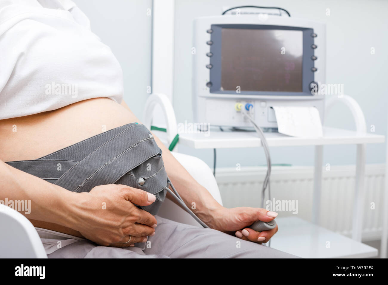 Pregnant woman sitting in clinic performing cardiotocography (CTG). The belt on her belly connected to the Cardiotocograph machine aka Electronic Feta Stock Photo