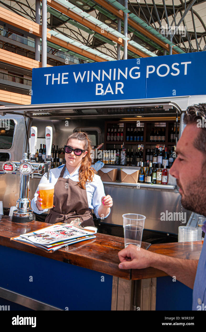 The Winning Post Bar at Royal Ascot racecourse, Berkshire, UK. Barmaid serving a customer with a draught pint of lager. Serving alcohol on sunny day Stock Photo
