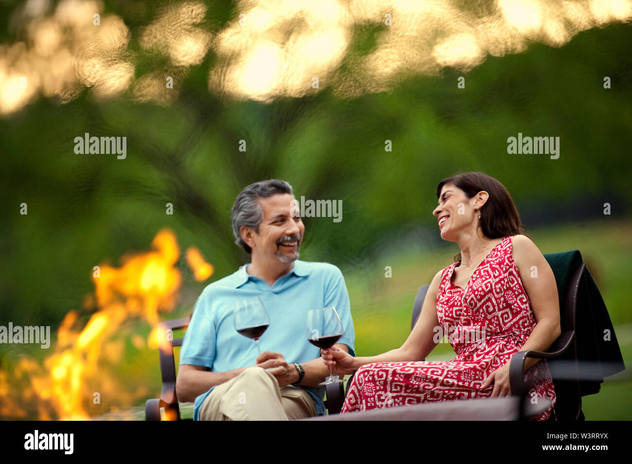 Smiling middle-aged couple relax with a glass of red wine beside a campfire in their garden in the late afternoon. Stock Photo
