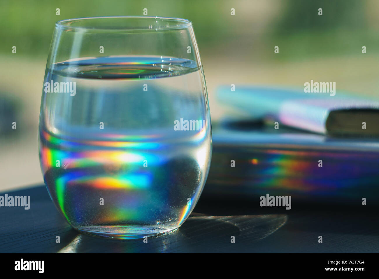Transparent glass filled with water reflecting rainbow spots from a notebook in silver holographic cover Stock Photo