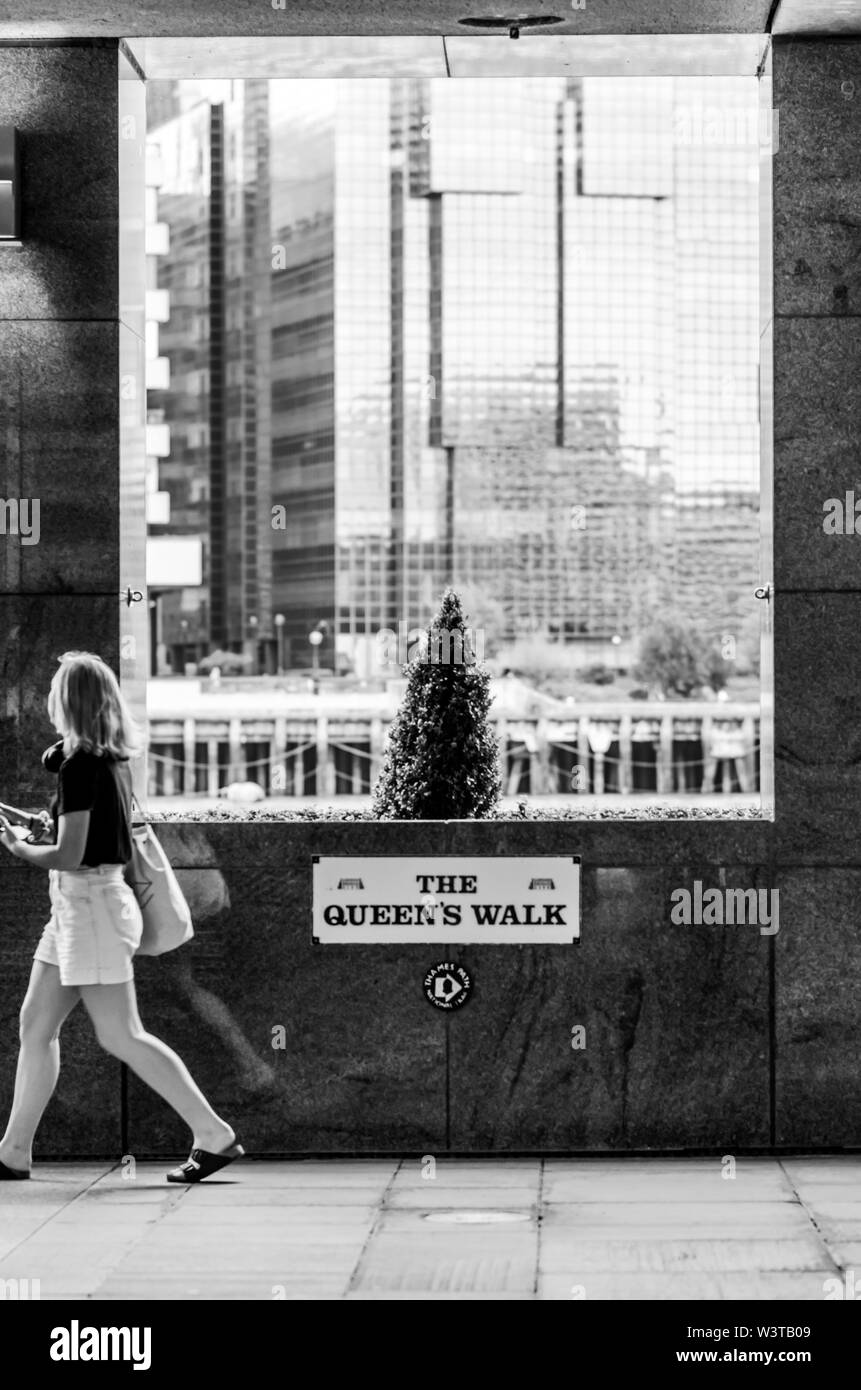 Black and white street photo of a young girl walking on The Queen's Walk promenade in London and passing by the large window with a small tree and an Stock Photo