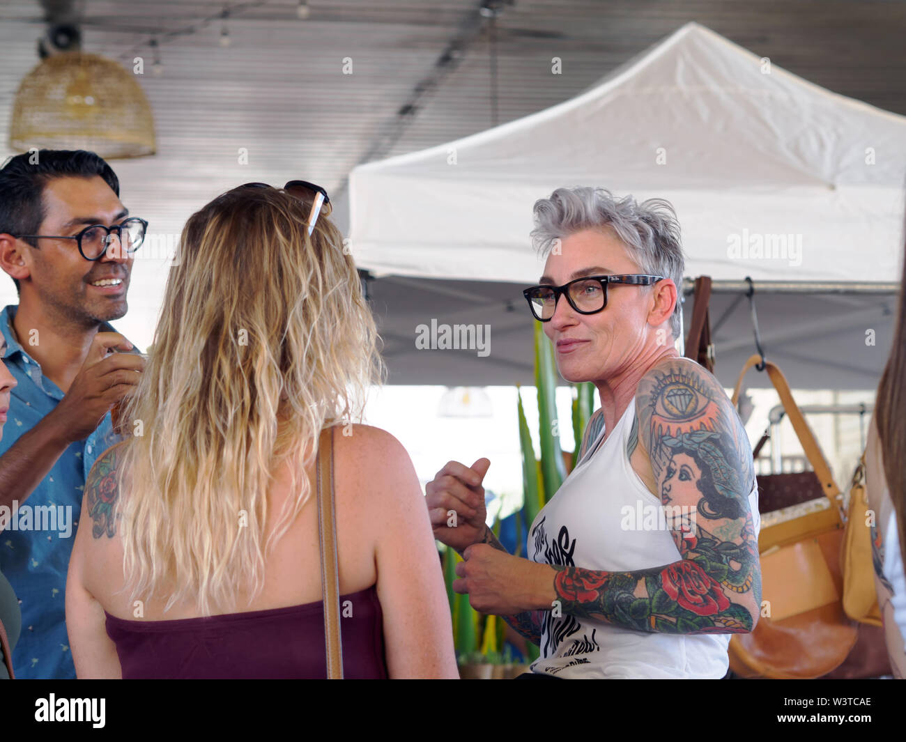 A fit, heavily tattooed mature woman in glasses and tank top converses with people at a 'Loca for Local' event in downtown Corpus Christi, Texas USA. Stock Photo