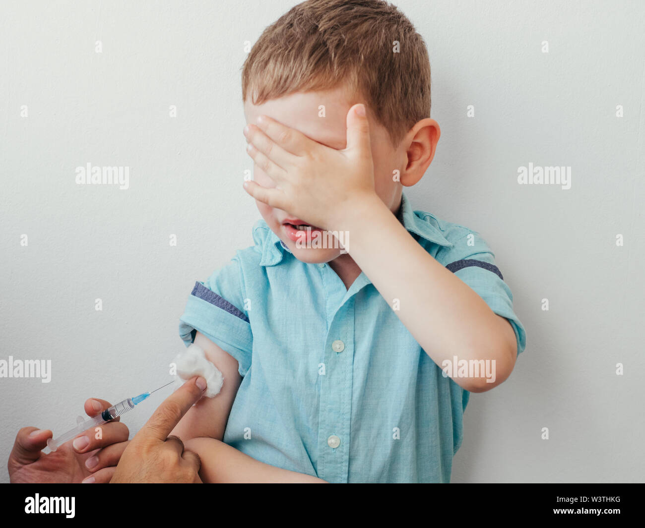 Little boy is afraid to vaccinate. Little caucasian baby boy in blue shirt covered his eyes with hand during injection. Vaccination of children concept. Stock Photo