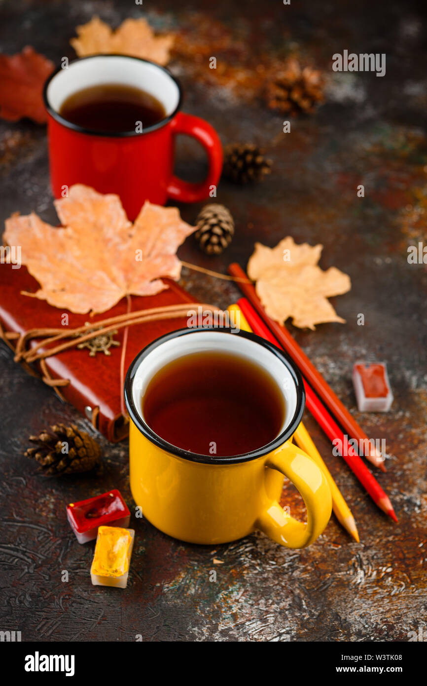 Yellow enamel cups of tea, watercolors in cuvettes, colored pencils, autumn maple leaves and bumps on a brown background Stock Photo