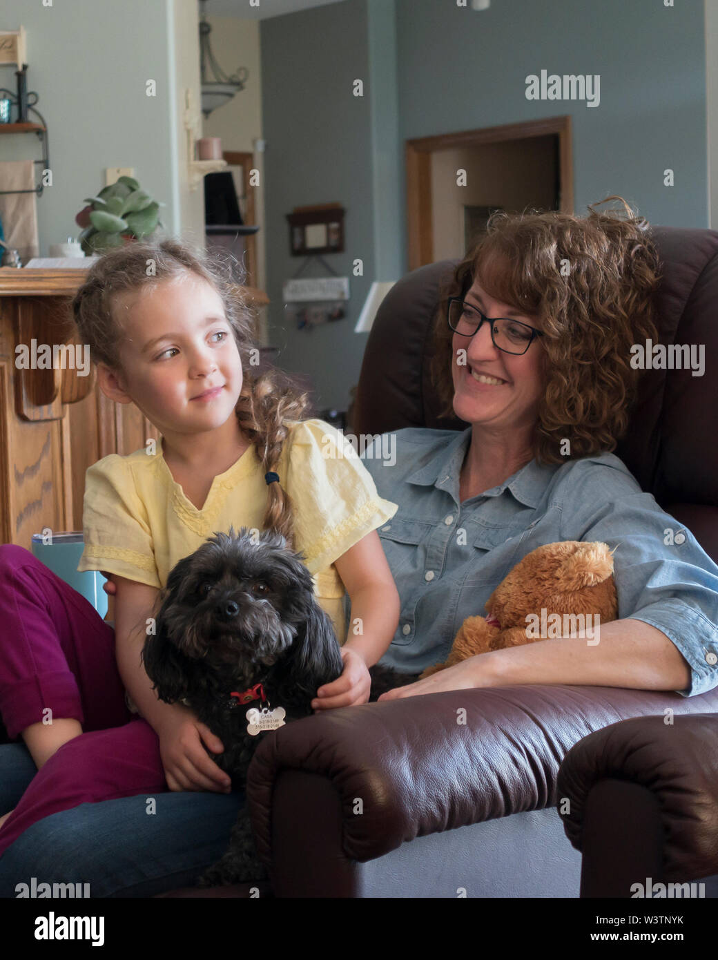 A smiling happy woman holds a five year old girl in her lap while the smiling child has her arms around a dog. Unposed image. USA Stock Photo