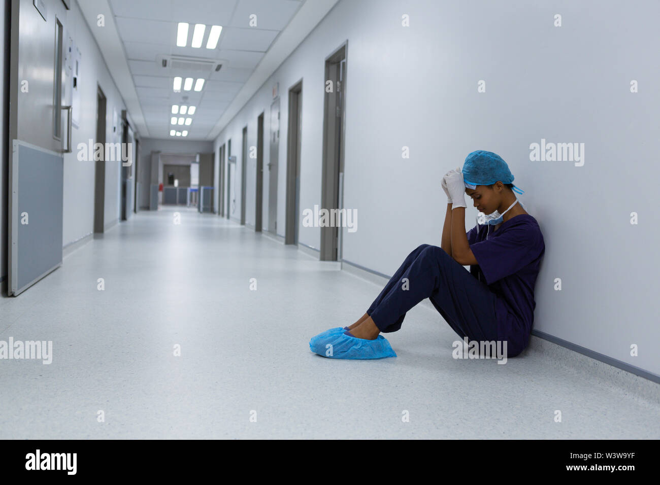 Tensed female surgeon with hands on forehead sitting on floor in the corridor at hospital Stock Photo