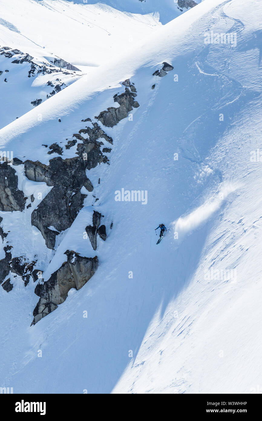 Skier drops into steep slope after several short radius turns. His skis throw snow from shadow into sunlight as he cuts close to the rocky cliffs abov Stock Photo