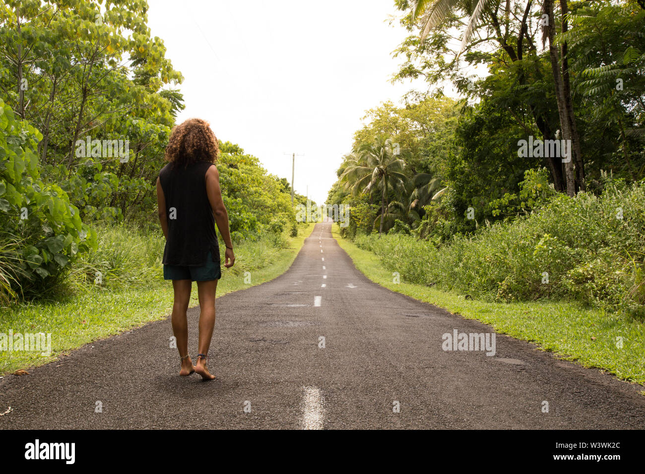Man, with curly hair, wearing black singlet, walking on empty road Stock Photo
