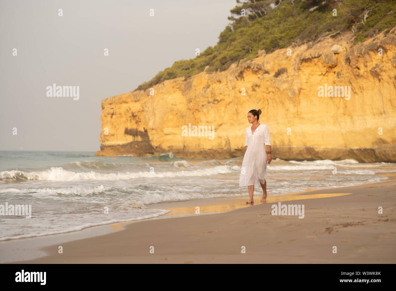 Woman with white dress walking along a wild beach with cliffs. Stock Photo