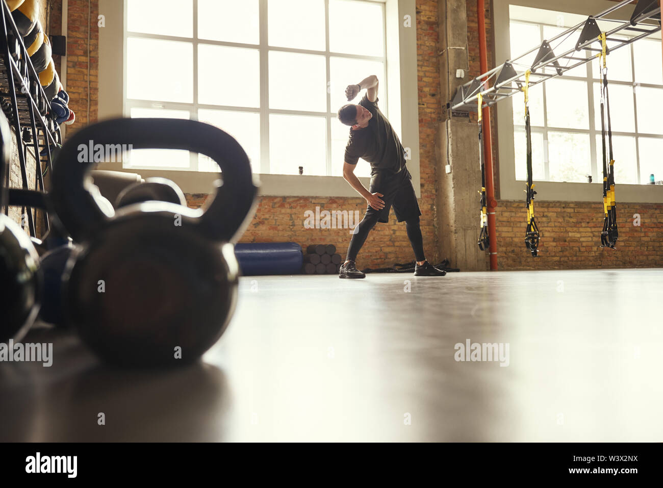 Warming up before training. Young sporty man in sportswear doing stretching exercises while standing in front of window at gym. Workout concept. Professional sport. TRX Training. Stock Photo