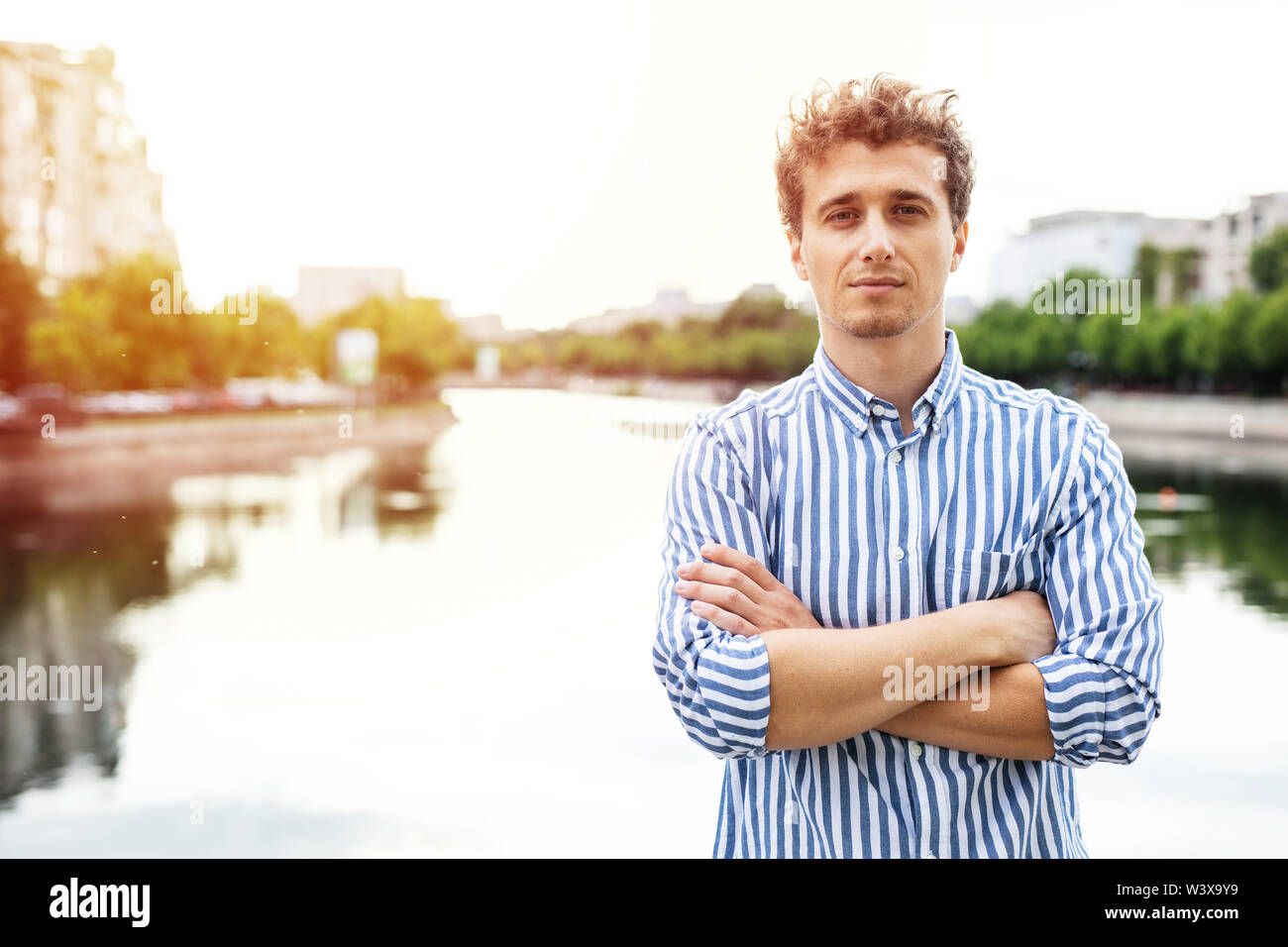 young man smart casual dressed looking with confidence to camera having a river behind, standing with arms crossed Stock Photo