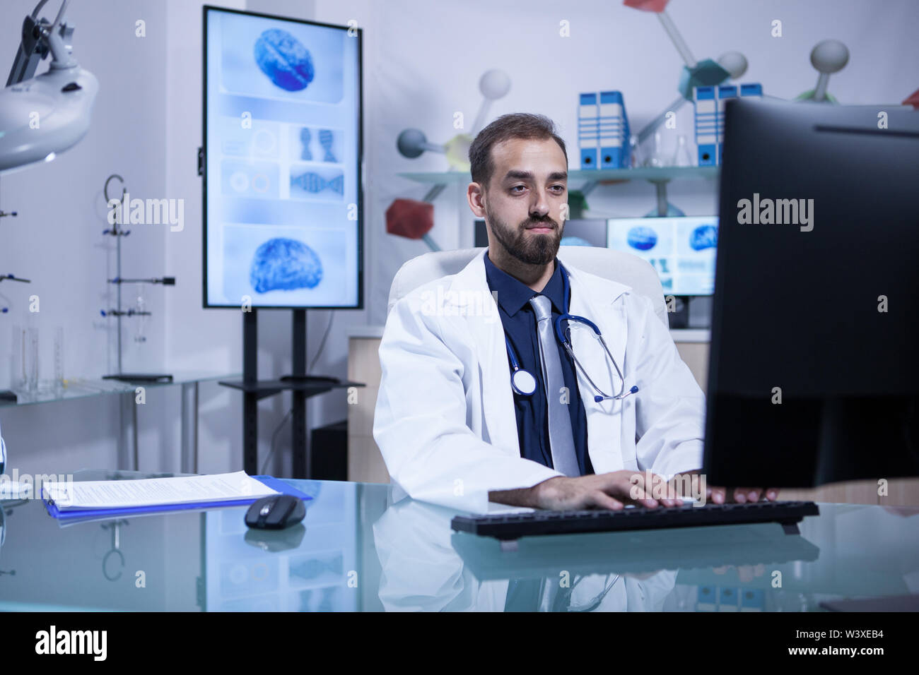 Young doctor siting at this computer typing a recipe for a patient. Doctor with stethoscope. Stock Photo