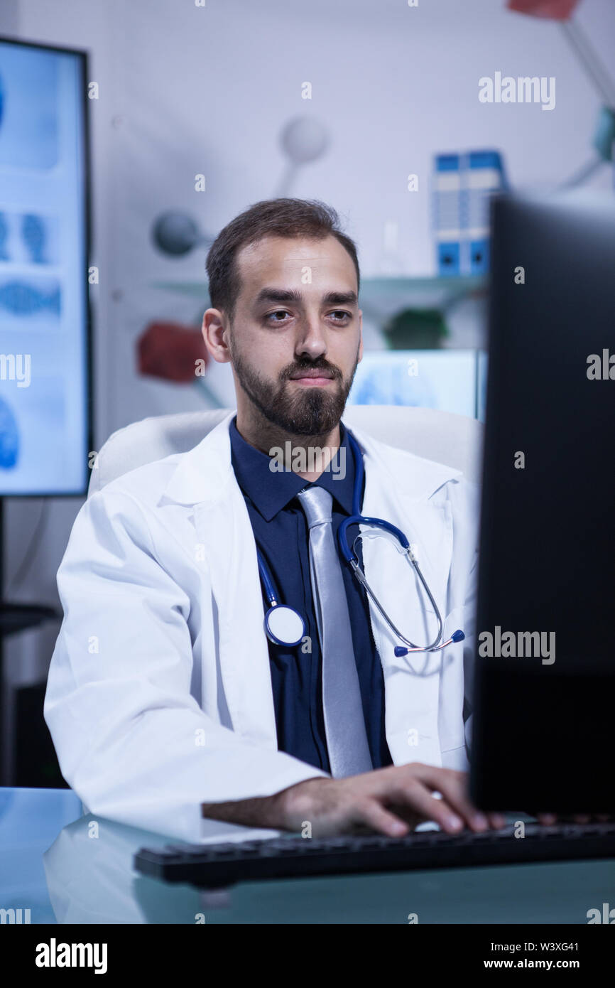 Handsome young bearded doctor typing in his computer in the hospital office. Doctor in white coat with a stethoscope. Stock Photo