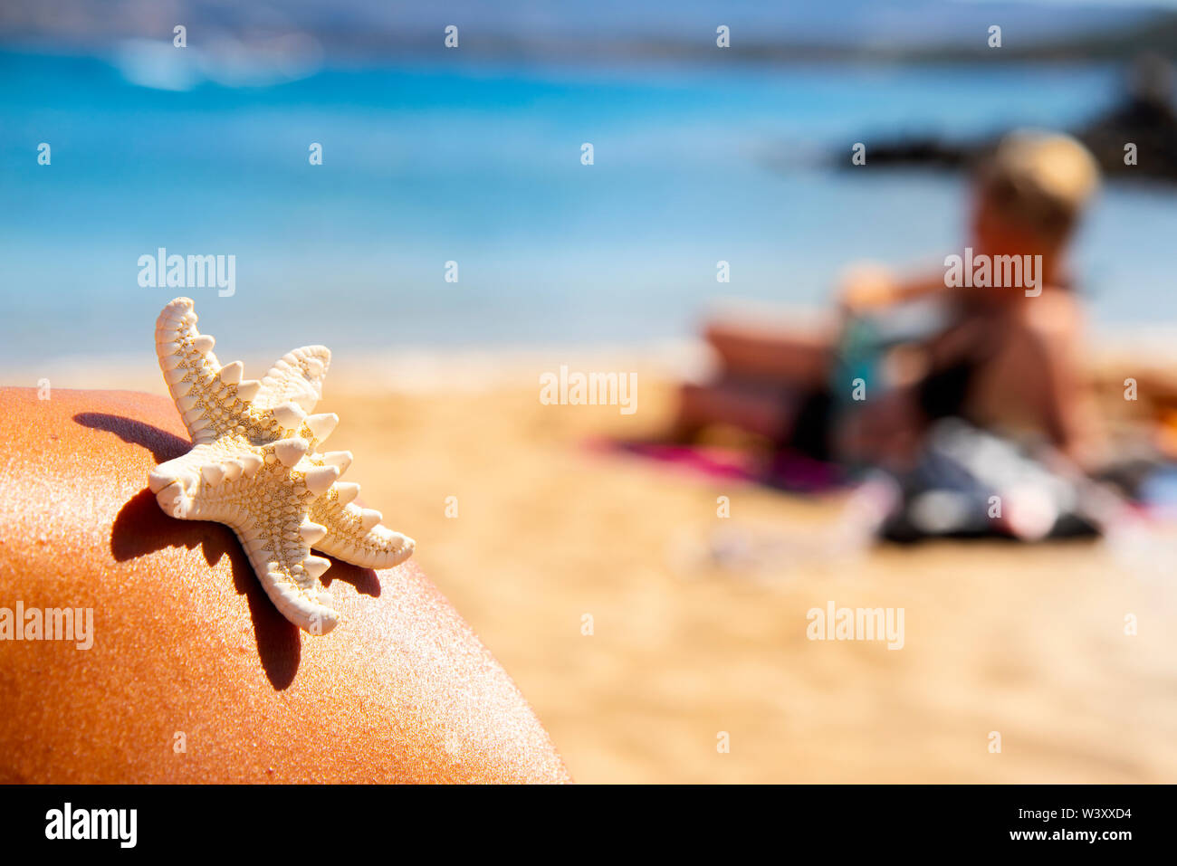 closeup of a white starfish on the shoulder of a young tanned person relaxing on the beach, with the sea and an unrecognizable person laying down in t Stock Photo