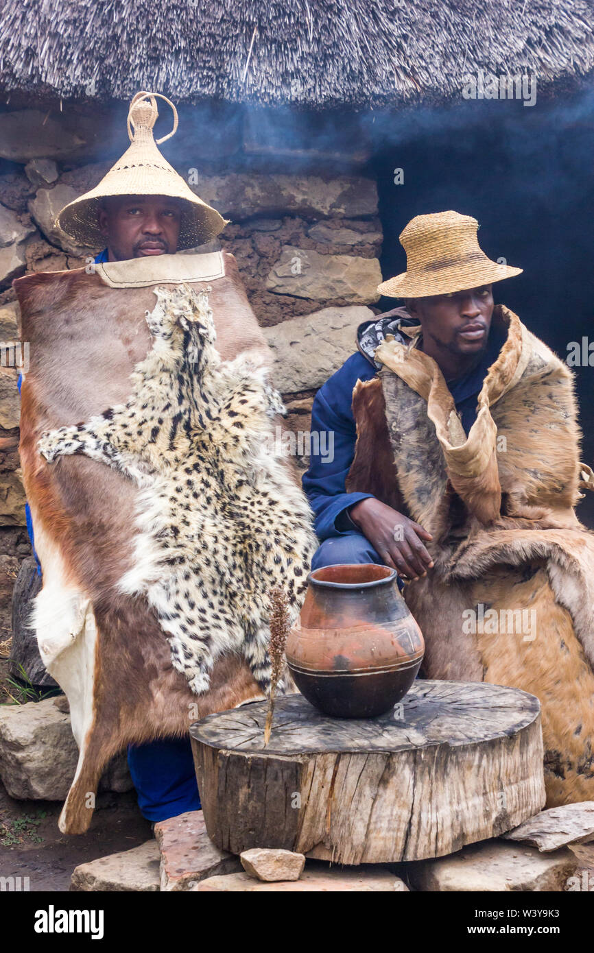 two Basotho black African men sitting in traditional animal skins or hides outside a stone thatch rondawel or hut with a clay pot filled with beer Stock Photo