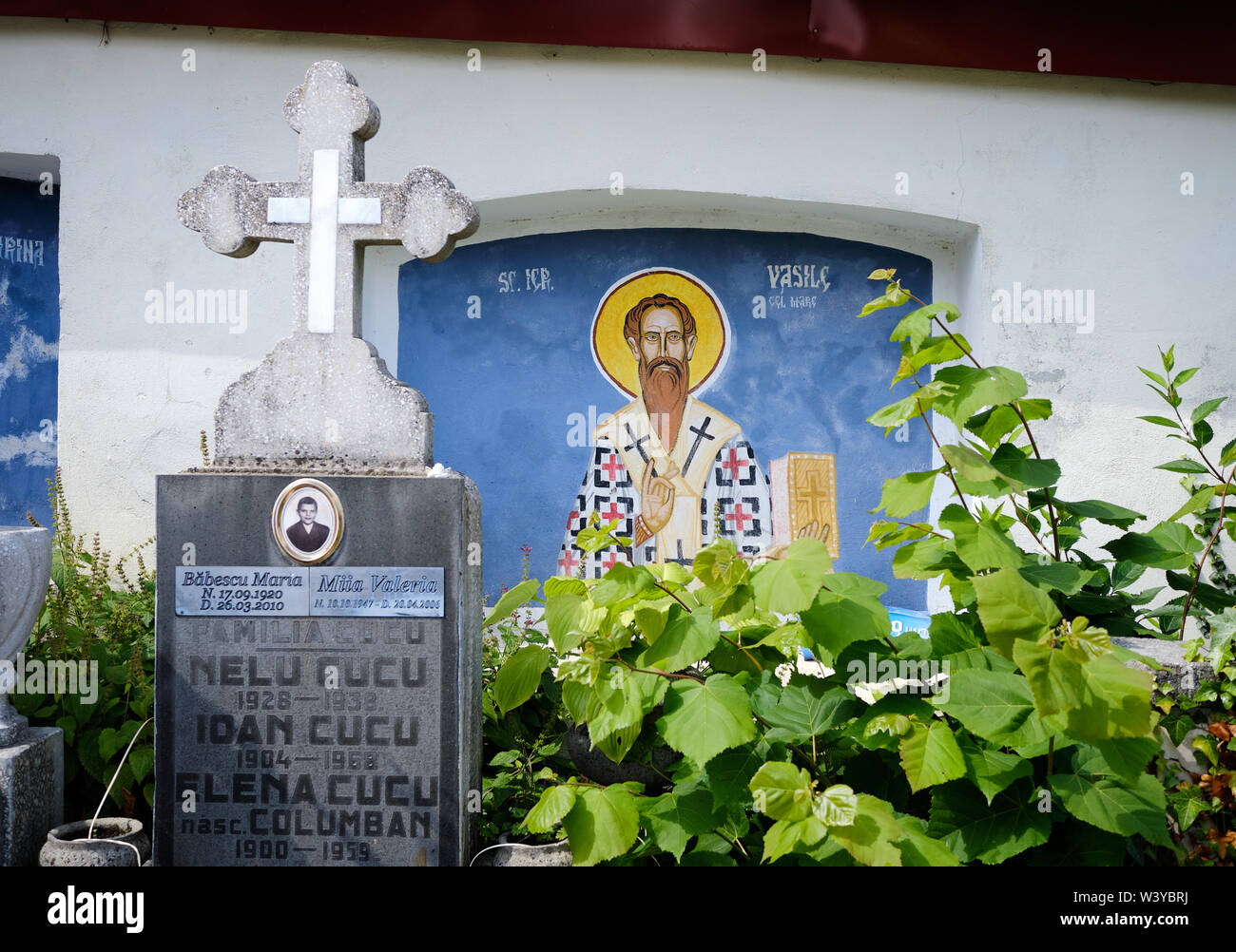 Painted wall facade with image of a saint in an Romanian Orthodox cemetery, framed by tombstone crosses and growing bushes Stock Photo