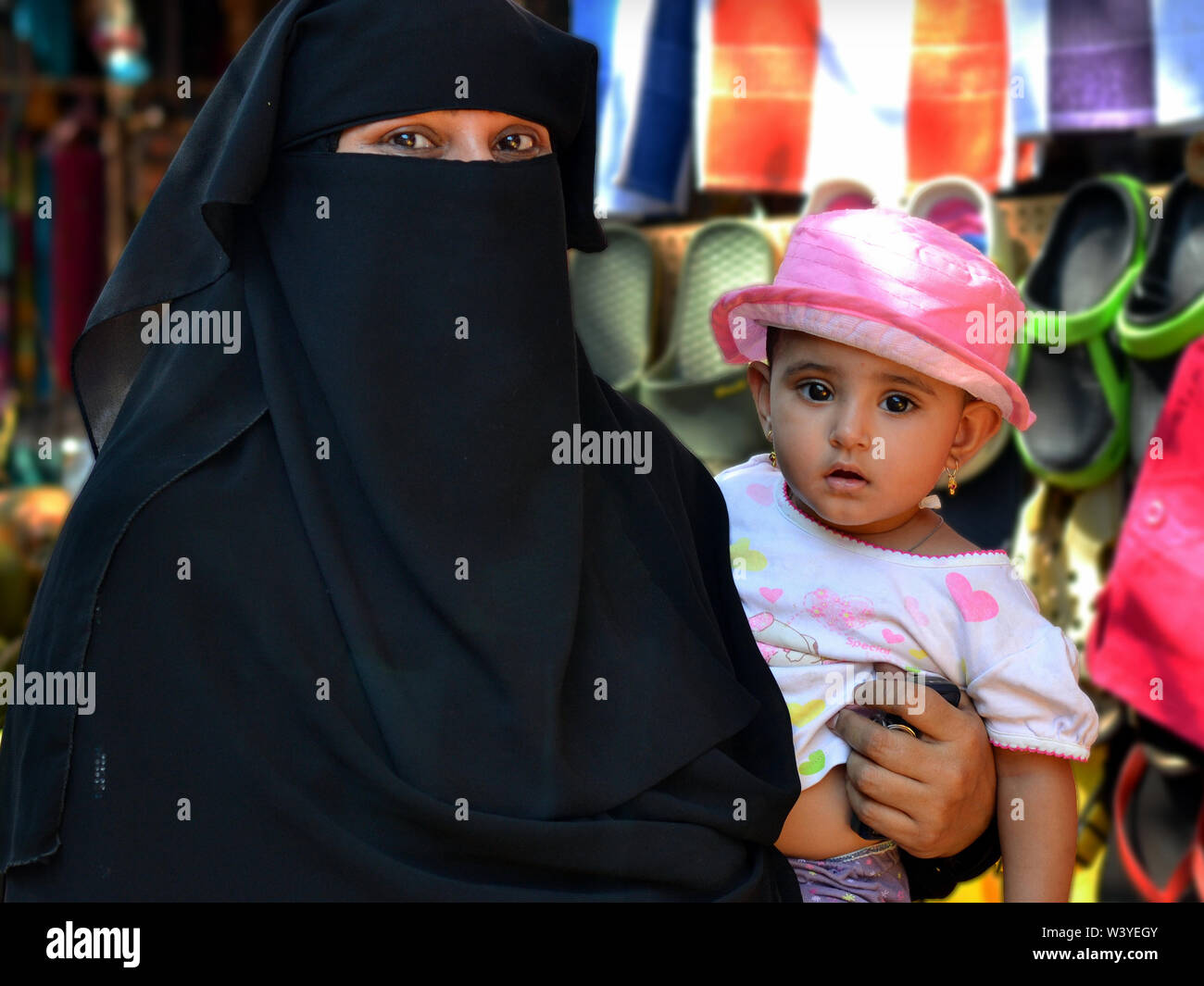 Indian Muslim woman with talkative eyes wears a black niqab and burqa and carries her little daughter in her arm. Stock Photo