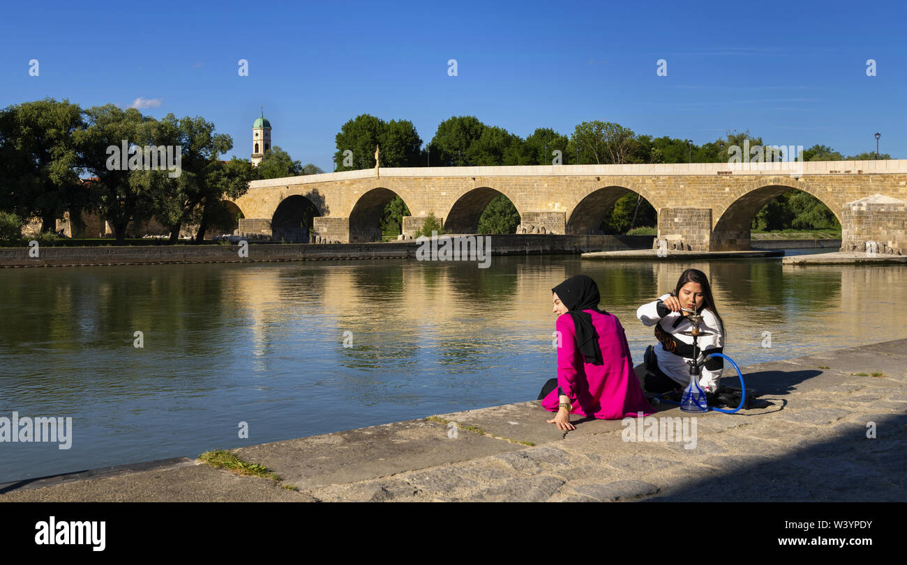 Regensburg, Germany, Juni 8, 2019: Two girls sit on the Danube embankment to smoke a hookah. In the backgroundis the historic stone bridge and the bew Stock Photo