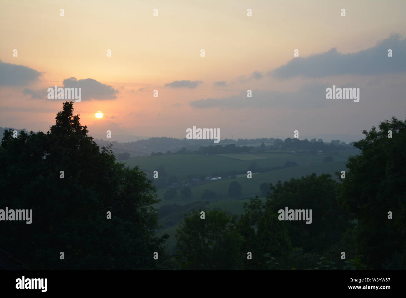 View of sun going down setting with trees in the foreground and hills to the distance re summer calm and cloudy early evenings South Herefordshire Stock Photo