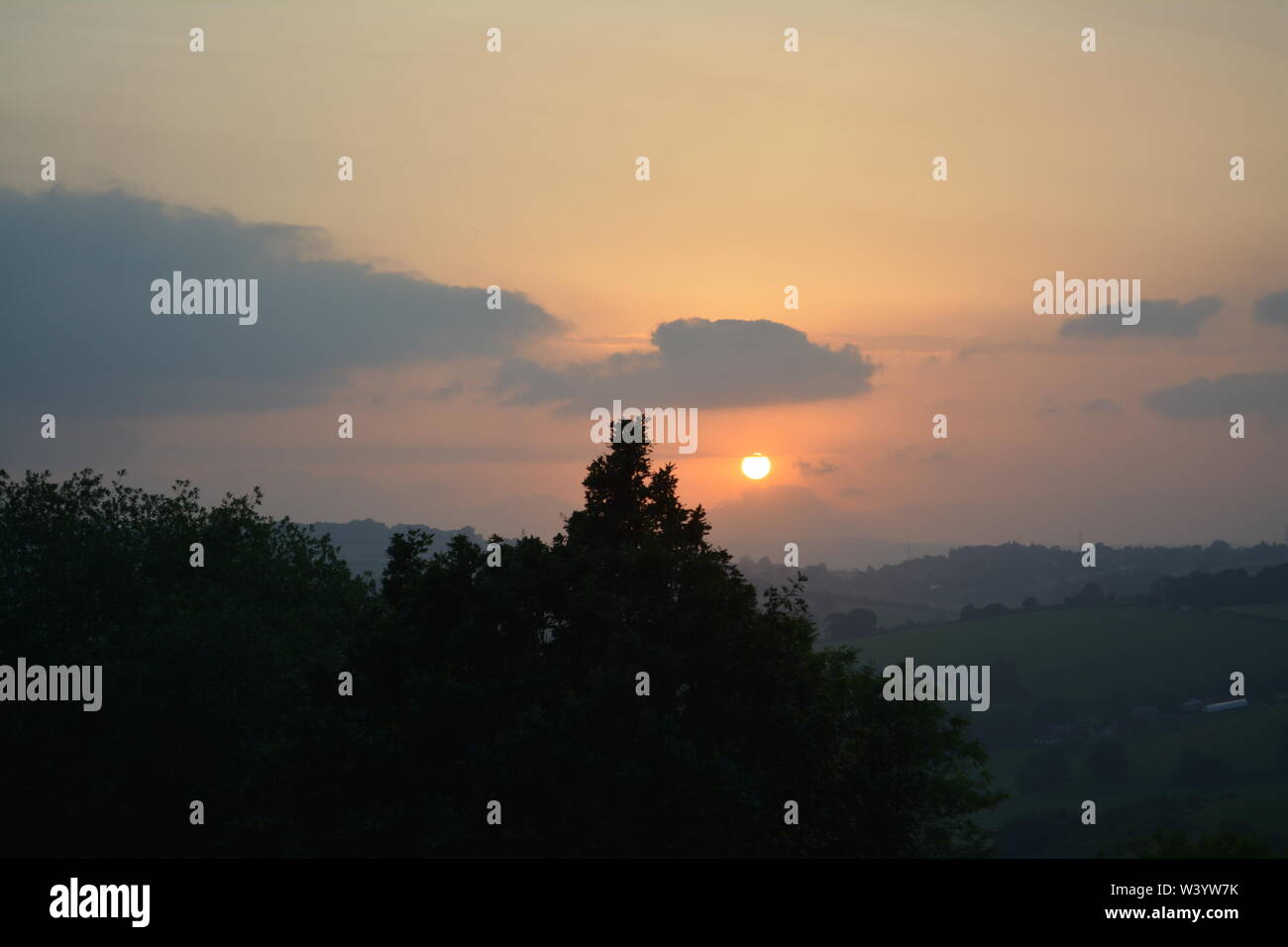 View of sun going down setting with trees in the foreground and hills to the distance re summer calm and cloudy early evenings South Herefordshire Stock Photo