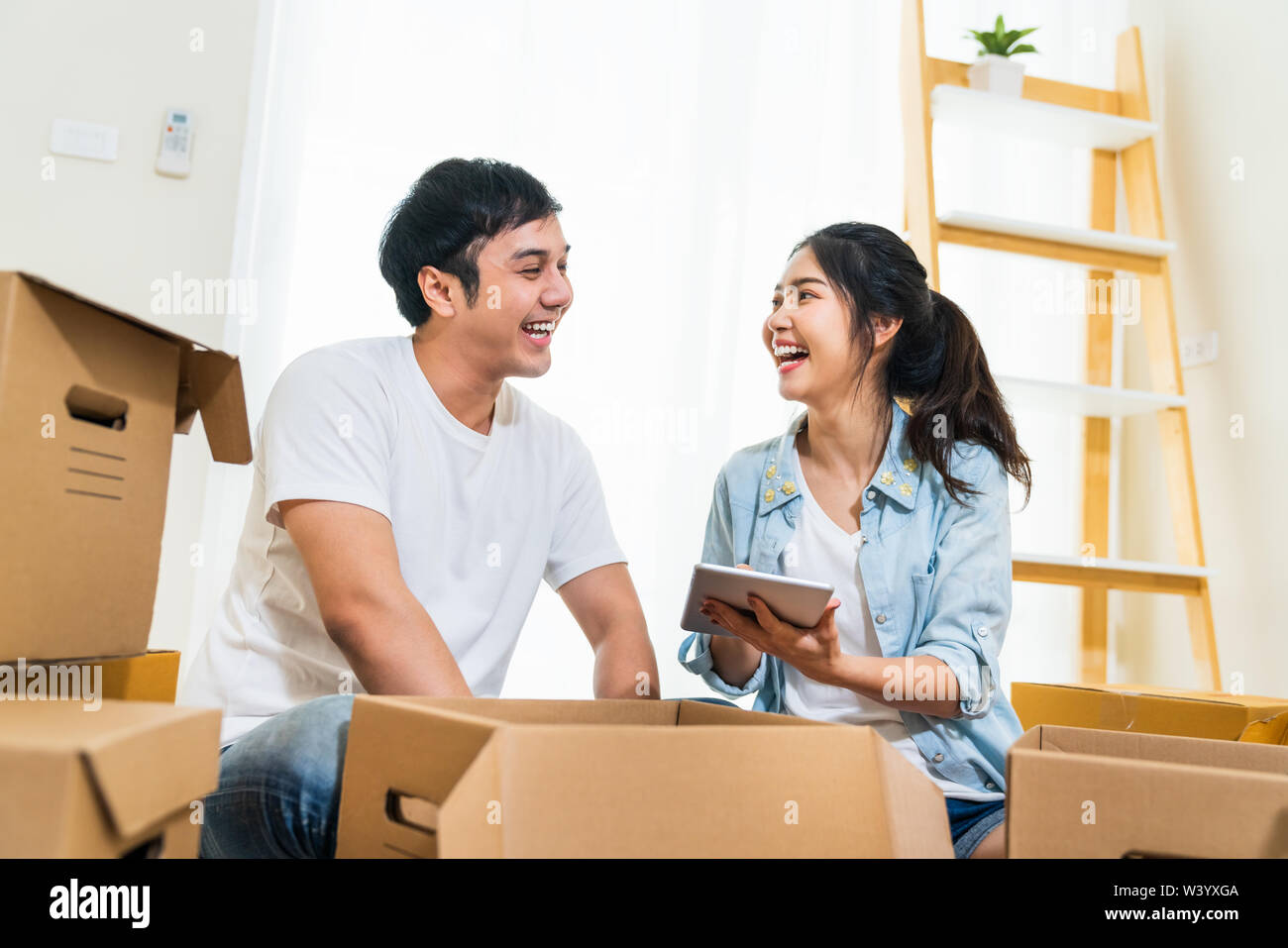 Happy young Asian couple moving in to new house, using digital tablet organizing things and unpacking boxes together. Home relocation, domestic life Stock Photo
