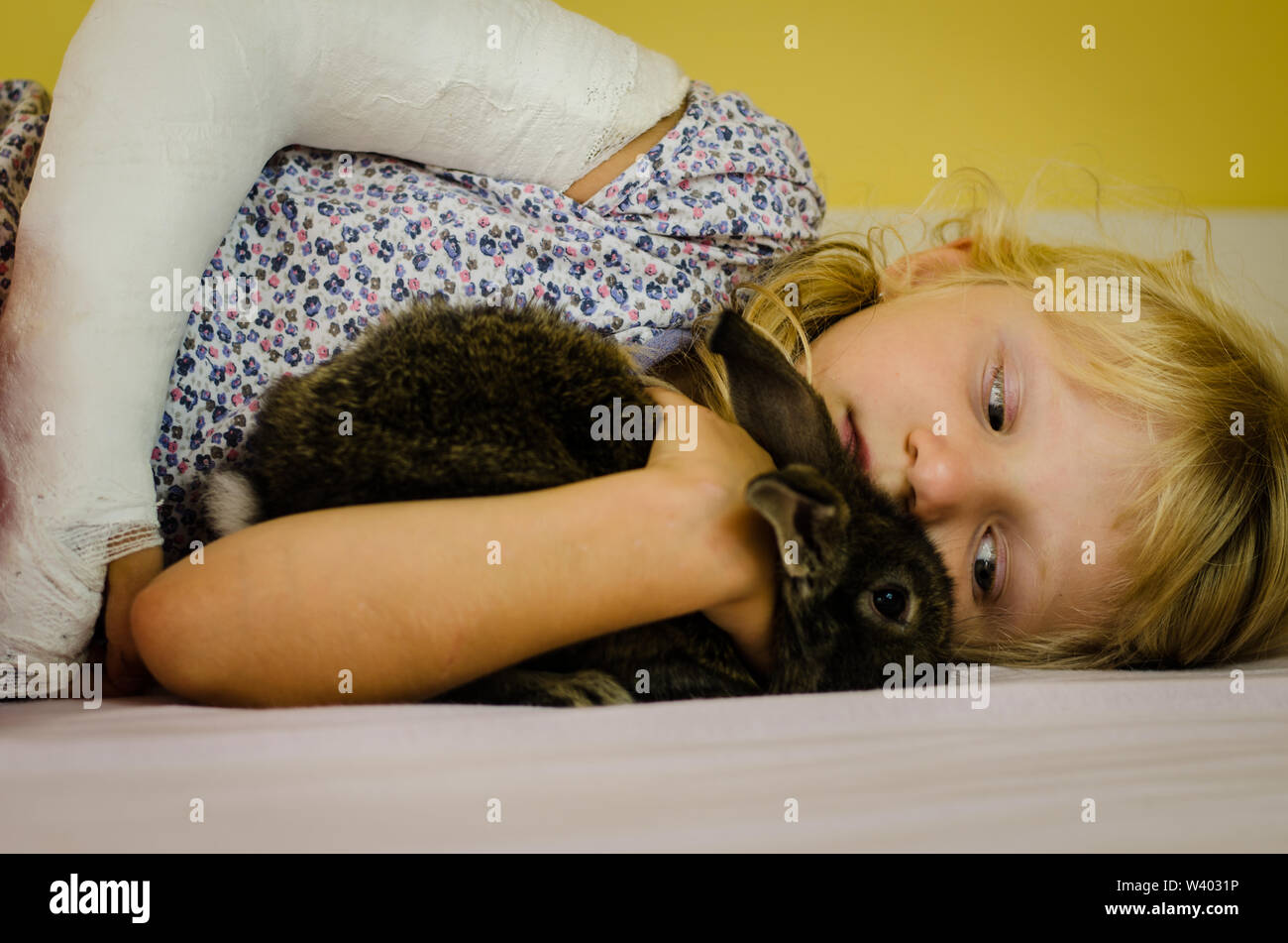 hurt blond girl with broken hand lying in bad and holding bunny rabbit pet Stock Photo