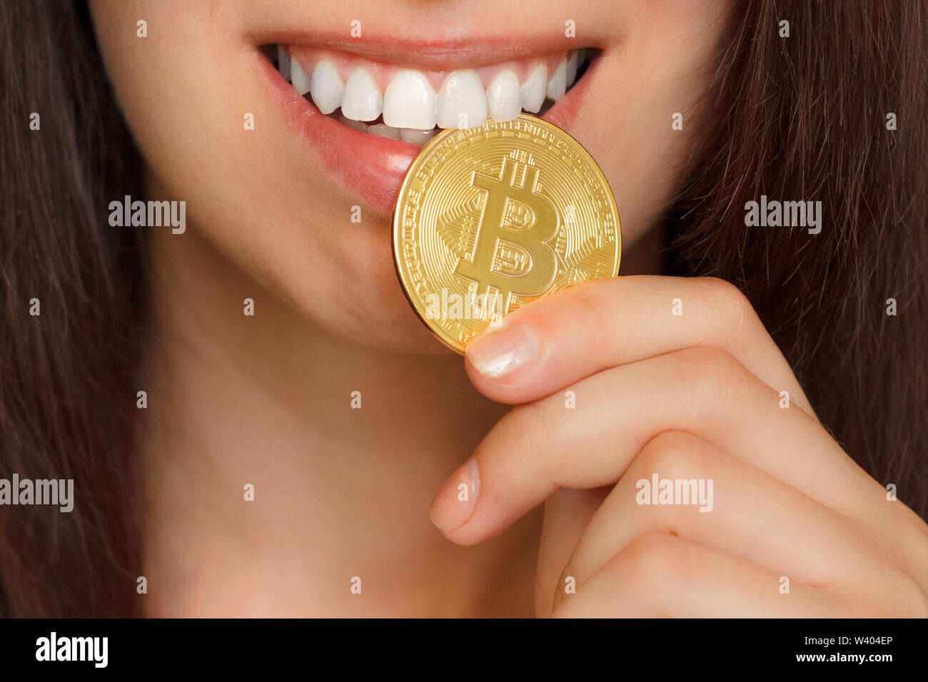 A young girl is biting of big golden BTC bitcoin by white teeth. Golden coin in the mouth. Stock Photo