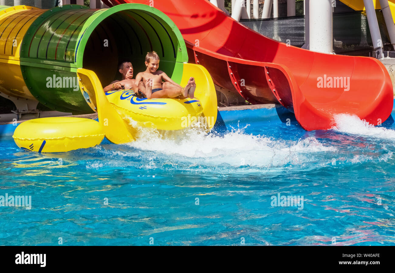 Family is riding down the water park structure, sitting together at inflatable ring and surrounded by water splashes. Father and son, adult and teenag Stock Photo