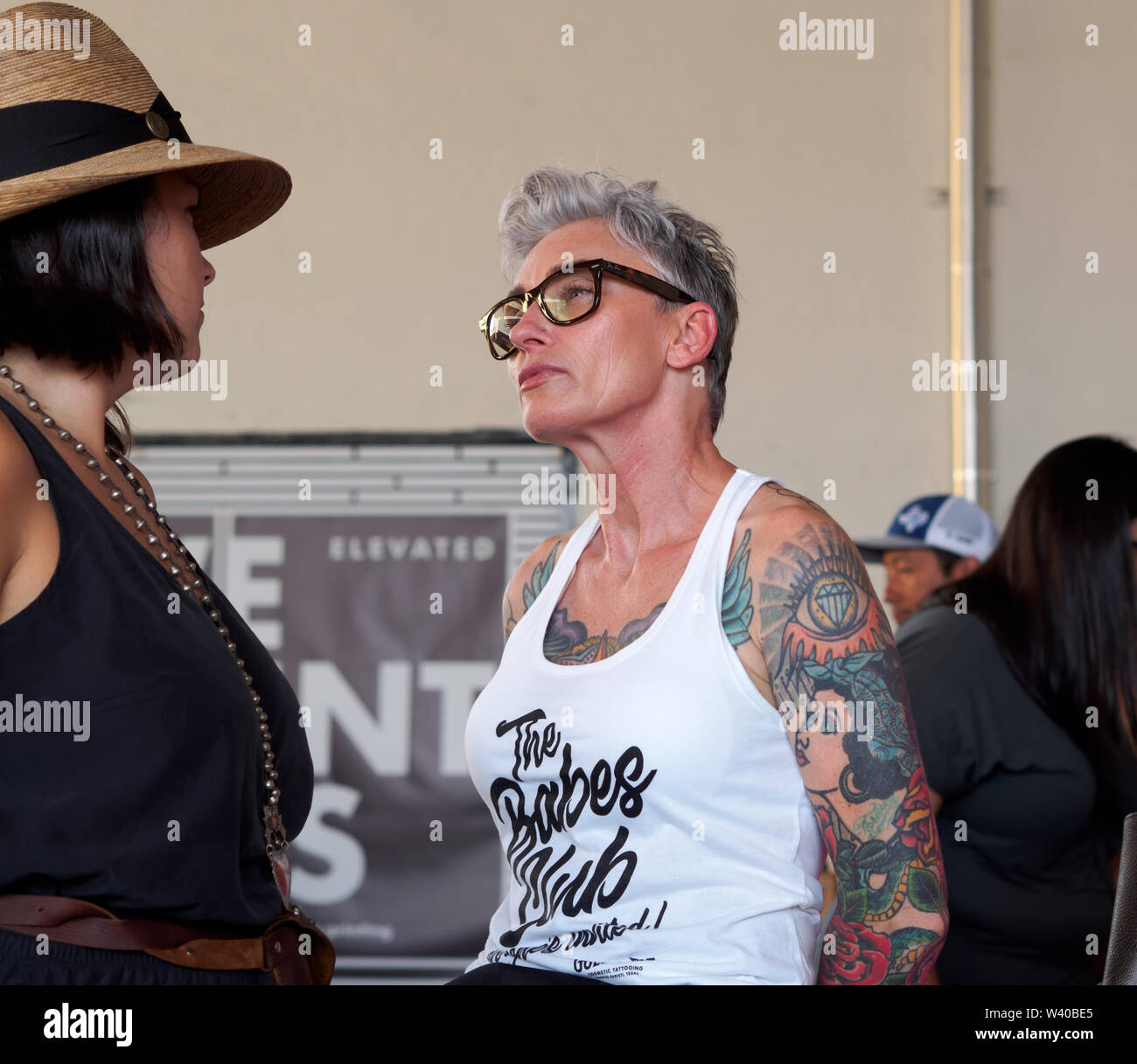 Fit, heavily tattooed mature woman in glasses and tank top converses with woman at a 'Loca for Local' event in downtown Corpus Christi, Texas USA. Stock Photo