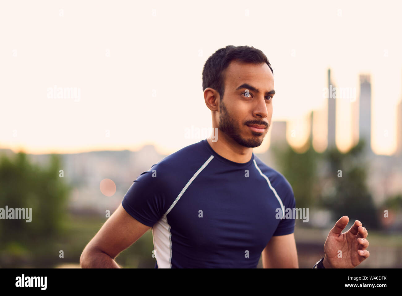 Portrait of active millenial man jogging at dusk with an urban cityscape and sunset in the background Stock Photo