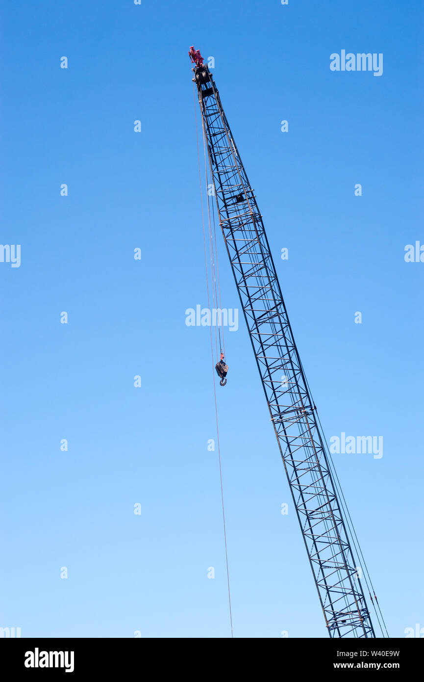 Tall industrial crane on clear blue sky, with cables and hook hanging from top pulleys. Stock Photo