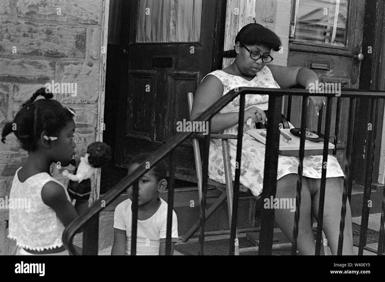African American 1960s mother and children, the girl is playing with a white doll that has black curly hair. Mother is listening to music on a portable record player she has on her lap some singles records, 45 rpm discs. They are on the porch of their home. New Brunswick, New Jersey,1969, USA 60s US HOMER SYKES Stock Photo