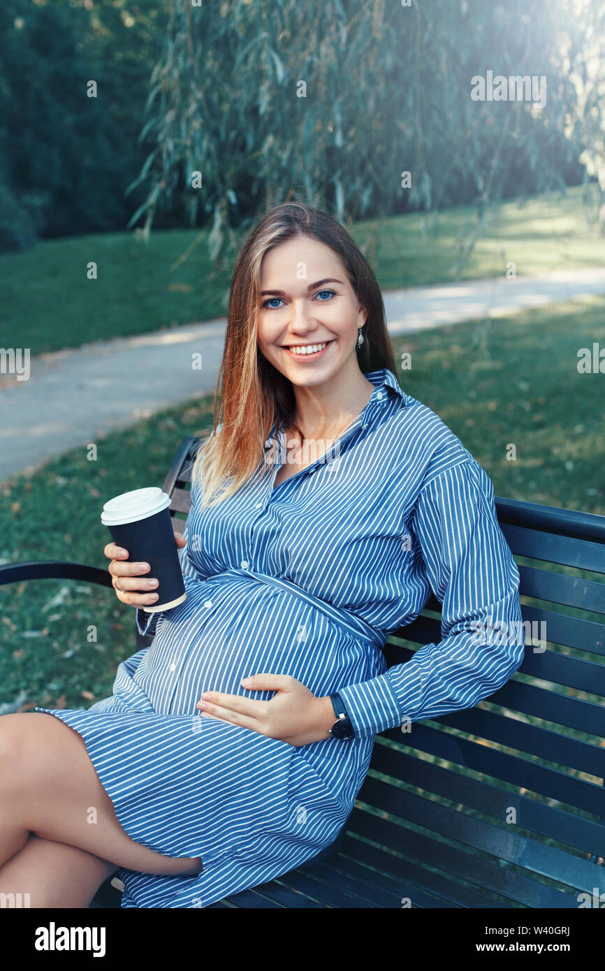 Happy healthy maternity. Portrait of pregnant young Caucasian woman sitting on bench in park outside and drinking coffee from paper cup. Unhealthy bad Stock Photo