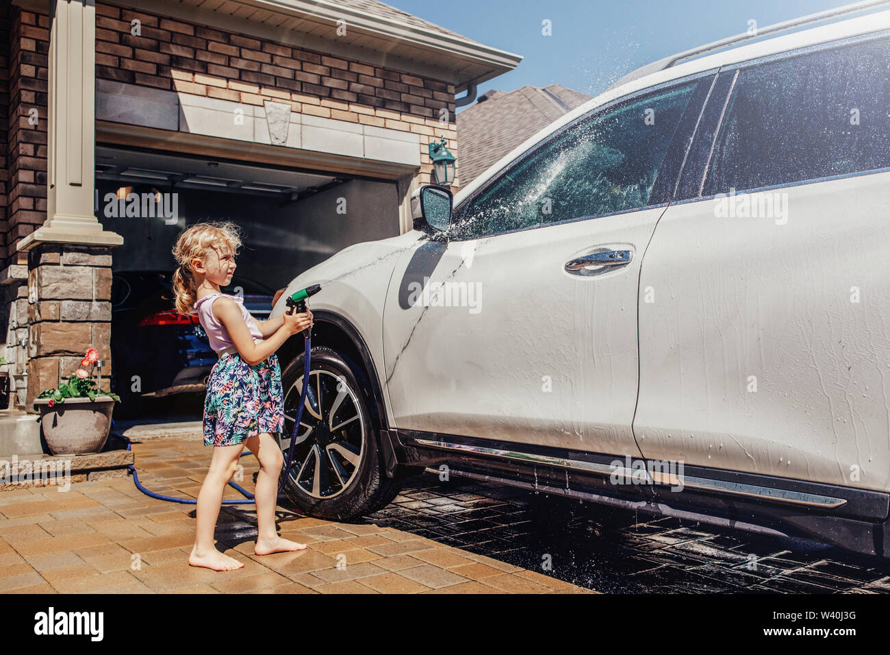 Cute preschool little Caucasian girl washing car on driveway in front house on sunny summer day. Kids home errands duty chores responsibility concept. Stock Photo