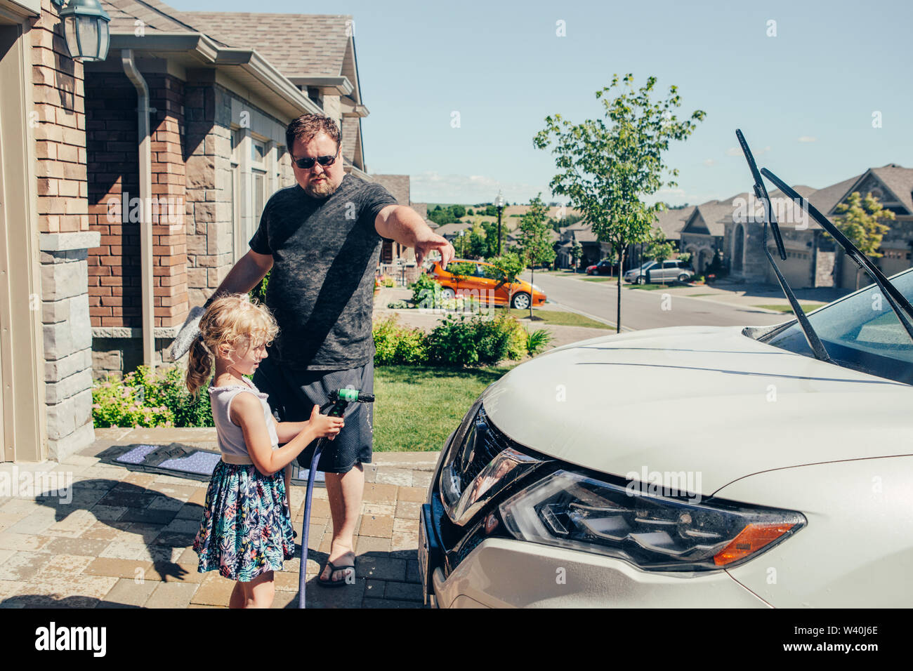 Cute preschool little Caucasian girl child helping father wash car with water from hose on driveway in front house on sunny summer day. Kids home erra Stock Photo