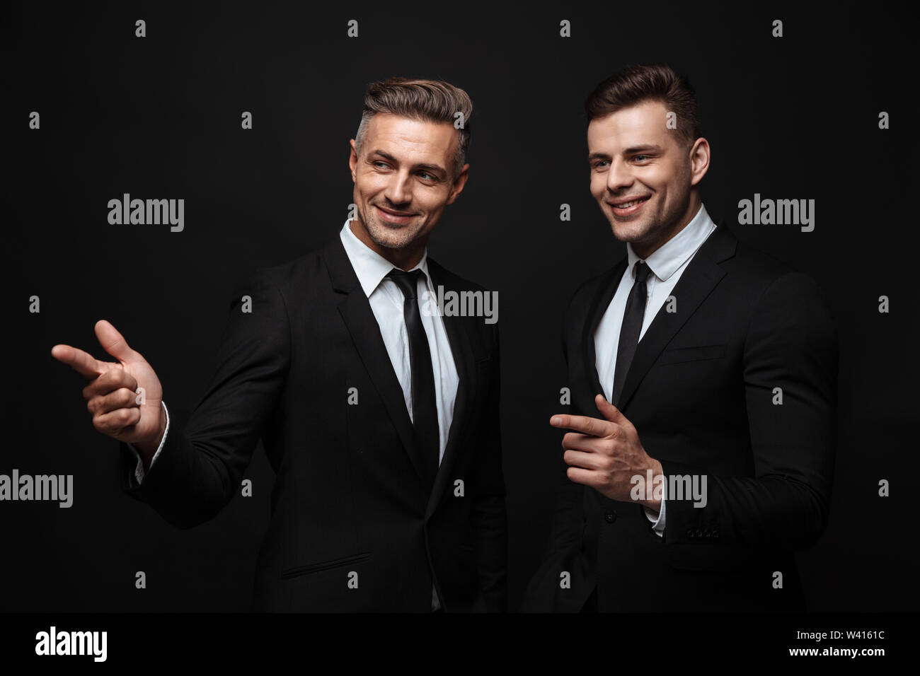 Portrait of smiling handsome two businessmen dressed in formal suit pointing fingers and looking aside isolated over black wall Stock Photo