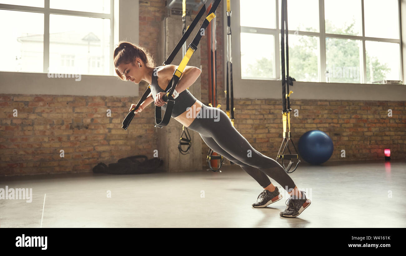 Working hard. Side view of young athletic woman with perfect body in sportswear doing push ups with trx fitness straps in the gym. Professional sport. TRX Training. Workout Stock Photo