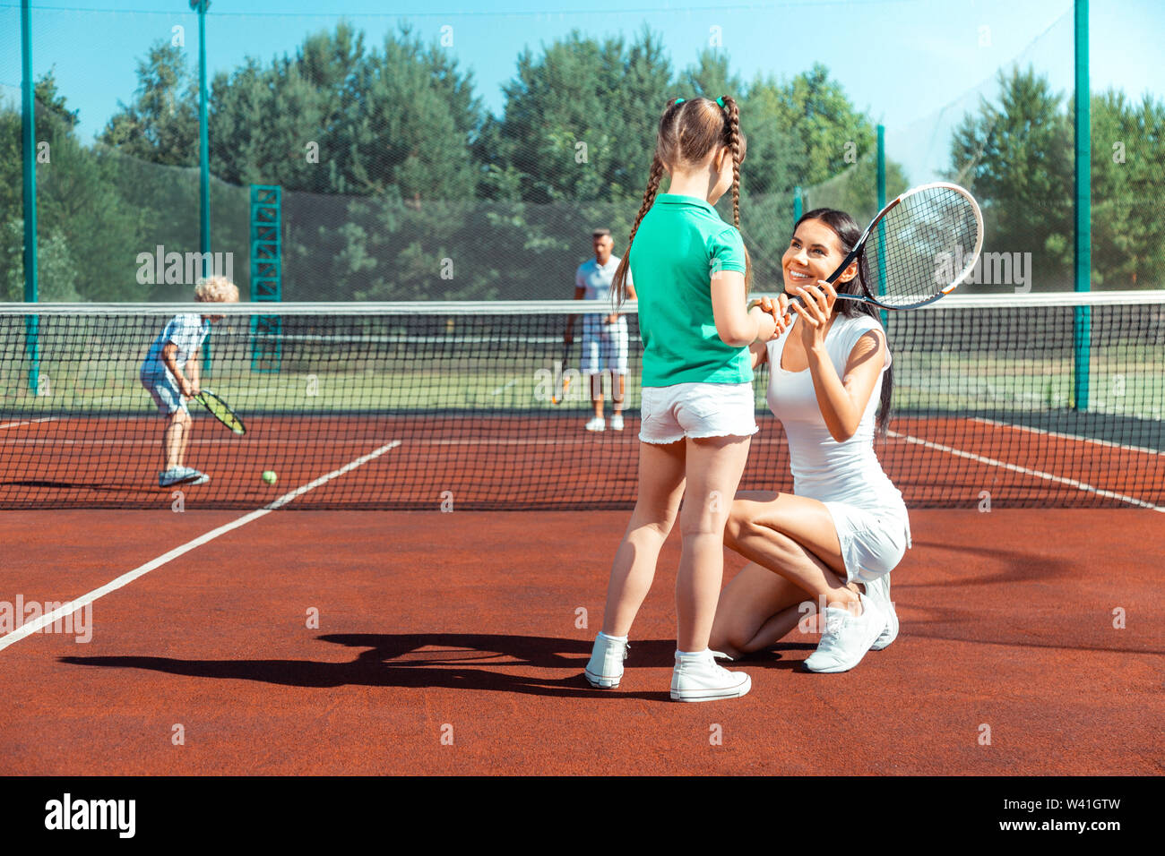 Dark-haired mommy smiling while teaching girl playing tennis Stock Photo
