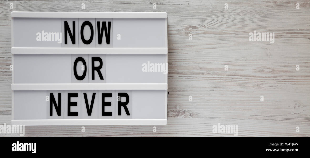 'Now or never' word on a lightbox on a white wooden background, top view. Copy space. Stock Photo