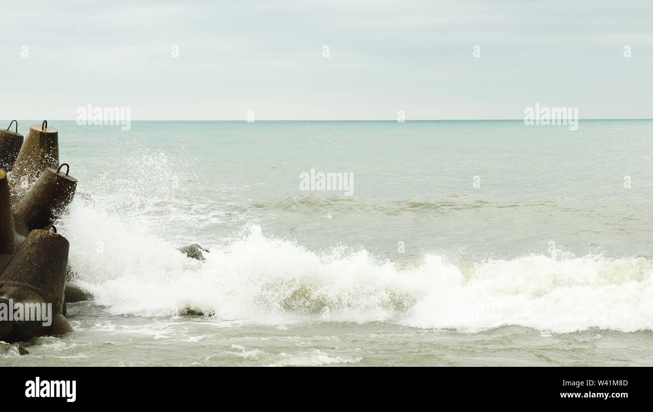 Waves break on shore. Bursts of seawater. Seashore before storm. Surges beat groyne. Dark cloudy sky over the sea. Water spray on shoreline. Stock Photo