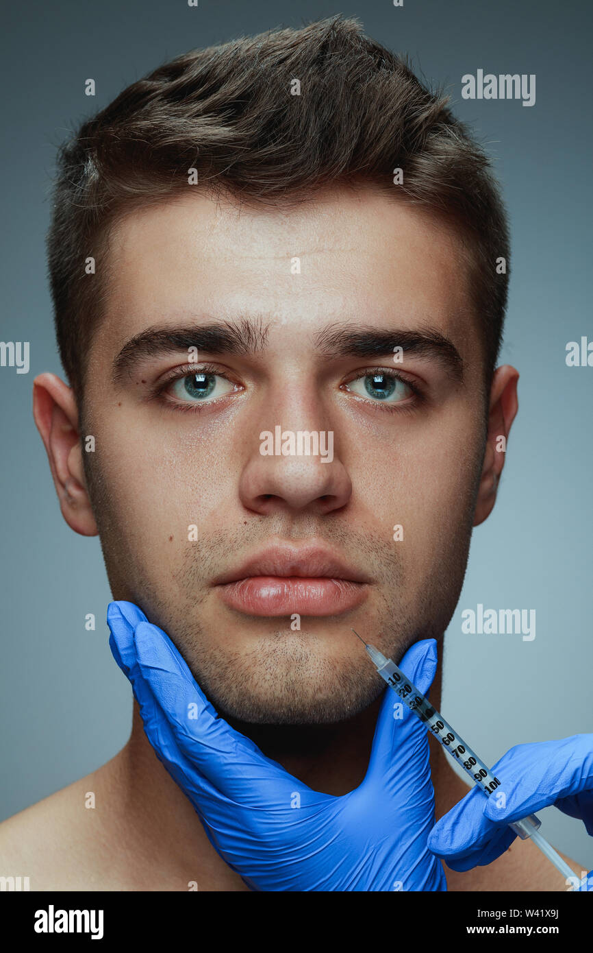 Close-up portrait of young man isolated on grey studio background. Filling botox surgery procedure. Concept of men's health and beauty, cosmetology, self-care, body and skin care. Anti-aging. Stock Photo