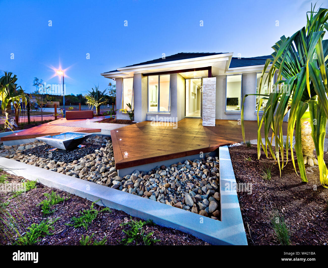 Modern mansion at night with lights on over the gravel garden with a pond near the pond beside the wooden floor, the house front included a tree Stock Photo