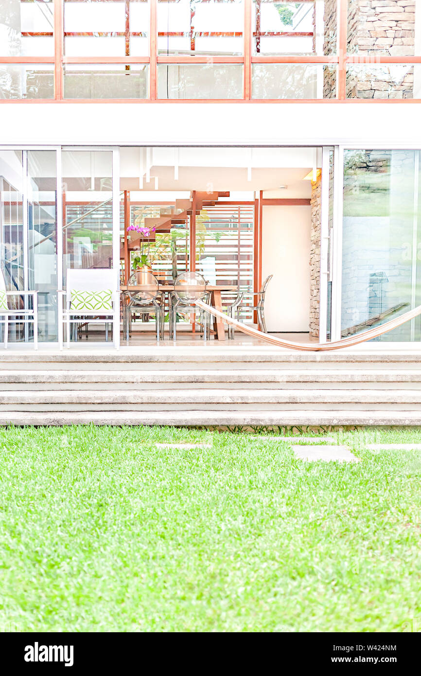 Modern house entrance with stone stairs and glass doors which showed chairs and tables inside through it, the green grass lawn near the chairs Stock Photo
