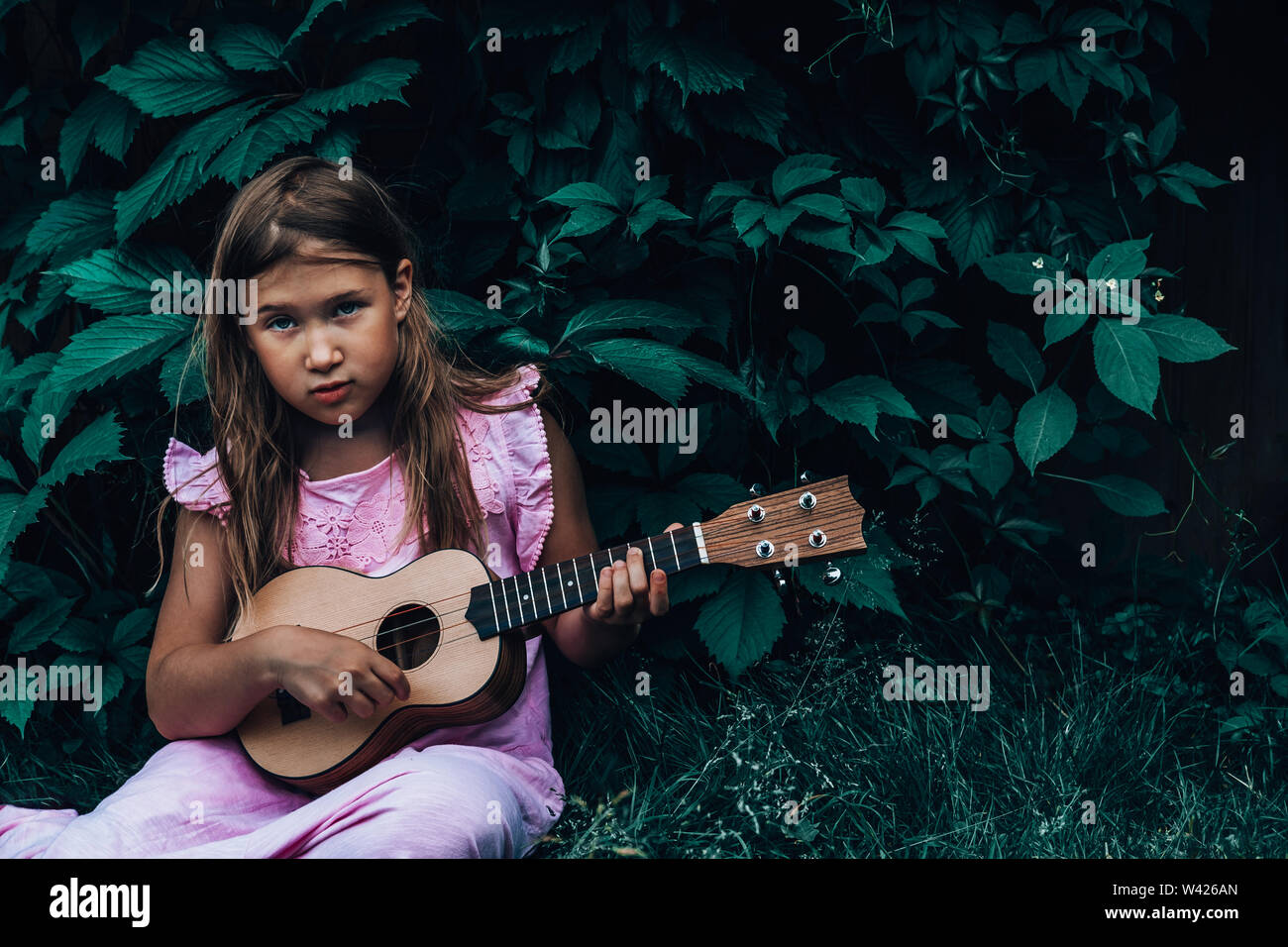 Beautiful young girl with blond hair and blue eyes wearing pink dress playing ukulele and dreaming on dark nature background. leaves. Cutie lady learns play concept Stock Photo - Alamy