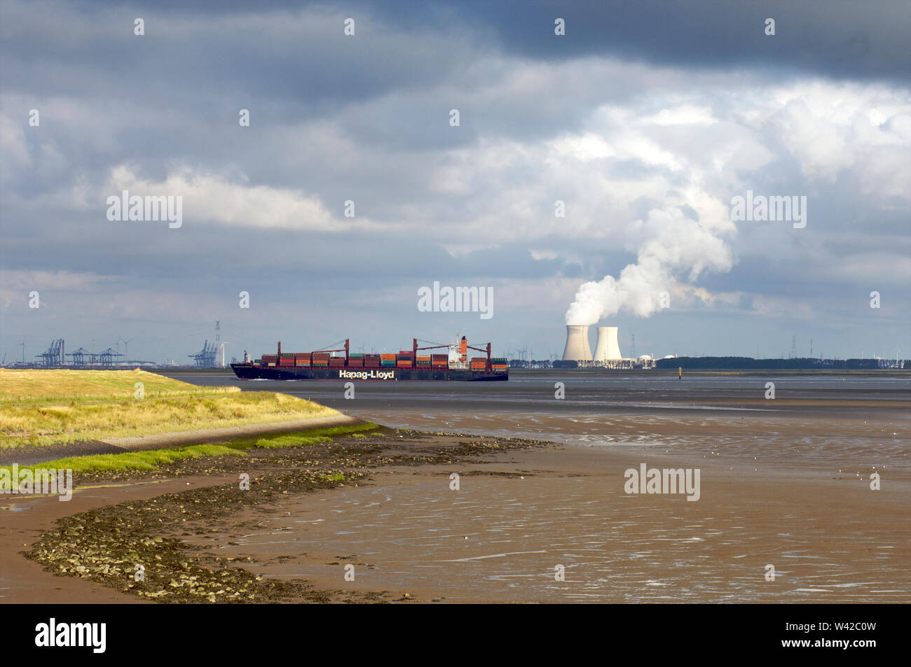 A container ship sailing over the Western Scheldt at low tide with the Doel nuclear power plant cooling towers in the background on Walcheren in Zeela Stock Photo