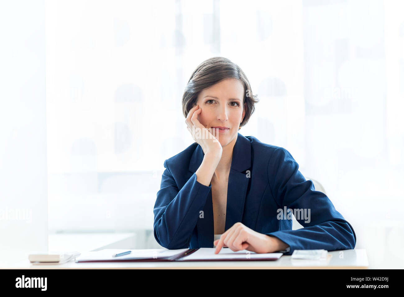 Thoughtful businesswoman staring pensively ahead at the camera with her chin resting on her hand as she sits at a desk in the office working on docume Stock Photo
